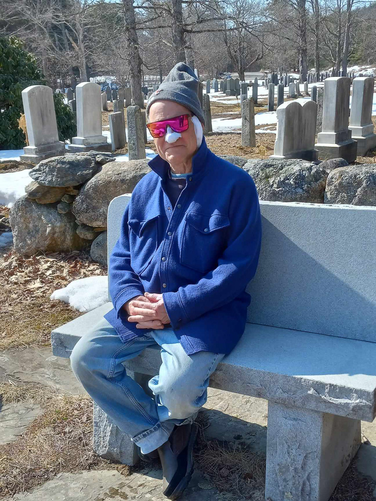 John sitting on a bench in a cemetery with a bandaged head